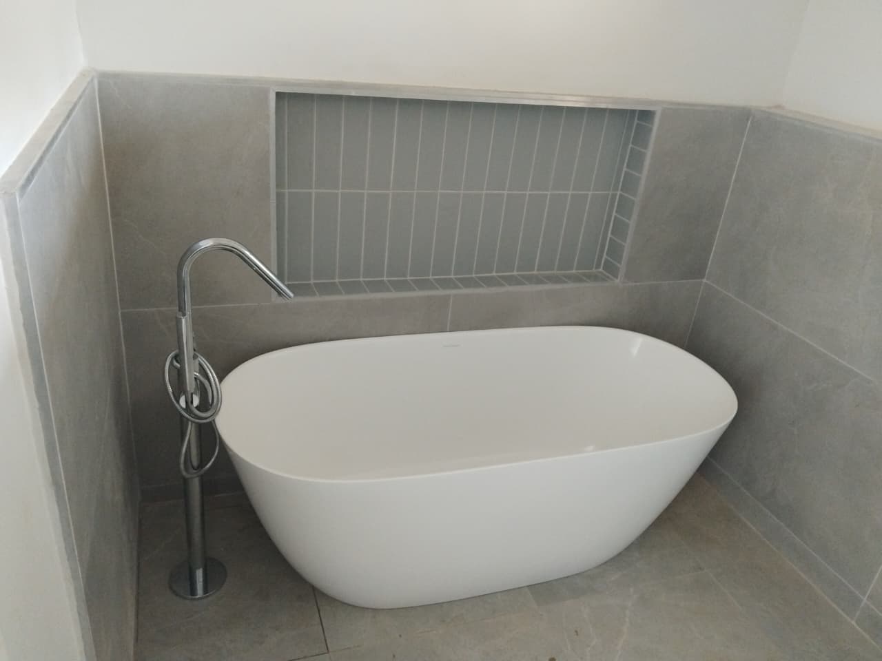 Freestanding bath installed against grey tiled walls with a floor-mounted mixer tap